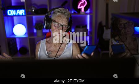 Technolo, une femme senior aux cheveux gris, passionne le monde du jeu vidéo en streaming, avec manette à la main, depuis sa salle de jeu éclairée par l'obscurité Banque D'Images