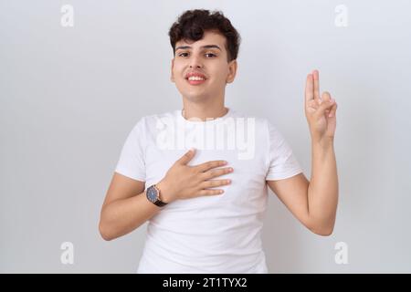 Jeune homme non binaire portant un t-shirt blanc décontracté souriant jurant avec la main sur la poitrine et les doigts vers le haut, faisant un serment de promesse de loyauté Banque D'Images