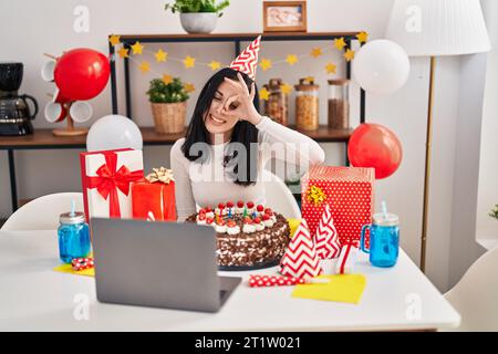 Femme hispanique célébrant l'anniversaire avec un gros gâteau au chocolat faisant appel vidéo souriant heureux faire ok signe avec la main sur l'œil regardant à travers les doigts Banque D'Images