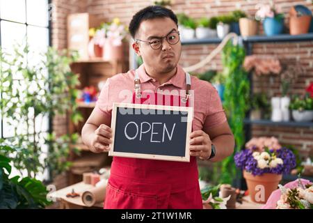 Jeune homme chinois travaillant au fleuriste tenant le signe ouvert sceptique et nerveux, fronçant les sourcils bouleversé à cause du problème. personne négative. Banque D'Images