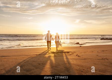 Couple romantique se promenant pieds nus sur la plage au coucher du soleil Banque D'Images