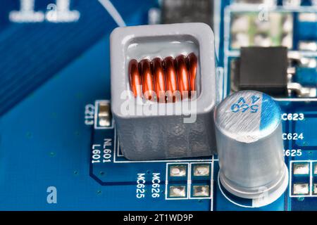 Gros plan de la bobine électronique et du condensateur électrolytique sur le fond de la carte de circuit imprimé bleue. Fil de cuivre de l'inducteur intégré en époxy dans un boîtier gris. Banque D'Images