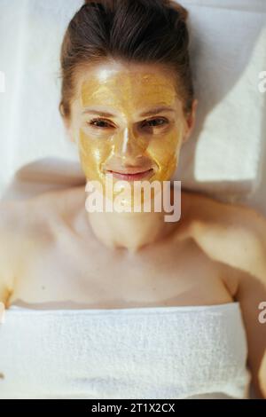 Le temps des soins de santé. Vue supérieure de la femme moderne détendue de 40 ans dans le salon de spa avec masque cosmétique doré sur le visage posé sur la table de massage. Banque D'Images