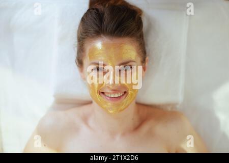 Le temps des soins de santé. Vue supérieure de femme moderne souriante dans le salon de spa avec masque cosmétique doré sur le visage posé sur la table de massage. Banque D'Images