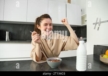 Portrait de jeune femme heureuse et rieuse mangeant des céréales avec du lait, triomphant, prenant le petit déjeuner et se sentant excitée, concept du matin énergique Banque D'Images