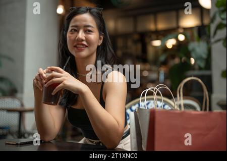 Une femme asiatique attrayante et confiante apprécie son café glacé tout en étant assise avec ses sacs dans un café ou un restaurant de la ville. Banque D'Images