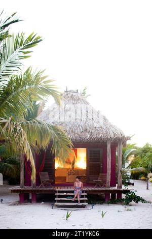 Une jeune femme est assise devant sa cabane de chaume dans une station balnéaire au Belize. Banque D'Images
