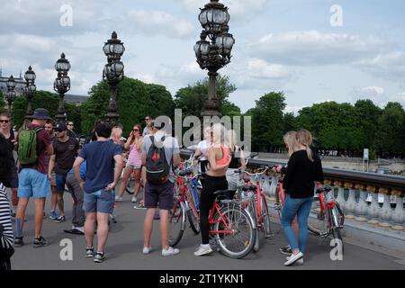 Un groupe de tour à vélo rencontre sur le pont Alexandre II le pont le plus orné de Paris, vélos, gens, lampes ornées ; un ciel nuageux et des arbres bordent la rivière Banque D'Images