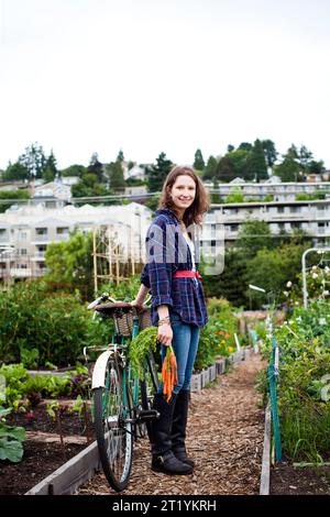 Une jeune femme sourit en regardant la caméra tout en se tenant à côté de son vélo et tenant un paquet de carottes. Elle est l'une des nombreuses personnes qui participent à l'agriculture urbaine et tendent quotidiennement t Banque D'Images