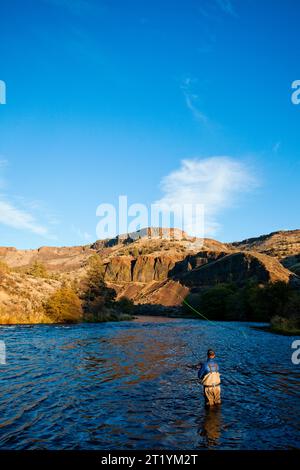 Un pêcheur à la mouche se tient dans l'eau profonde de la taille sur la rivière Deschutes avec des montagnes au loin sur une soirée ensoleillée à l'extérieur de Bend, Oregon. Banque D'Images