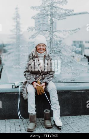 Femme senior assise à la glace de noël rinke dans la ville et puttion sur des patins à glace blancs. Banque D'Images