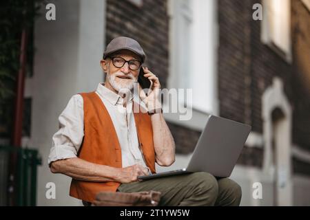 Homme âgé assis sur le trottoir de la rue travaillant sur un ordinateur portable, en utilisant les technologies numériques, faire un appel avec smartphone. Concept de seniors et compétences numériques. Banque D'Images