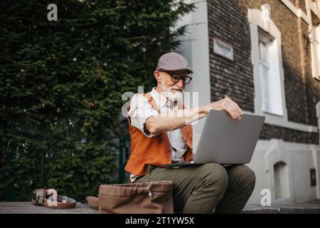 Homme âgé assis sur le trottoir de la rue travaillant sur un ordinateur portable, en utilisant les technologies numériques. Concept de seniors et compétences numériques. Banque D'Images