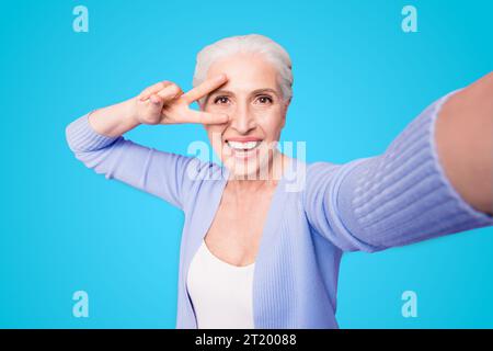 Une dame âgée fait un selfie sur l'appareil photo d'un smartphone faisant un v-sign près des yeux isolés sur fond violet clair avec un espace de copie pour le texte Banque D'Images