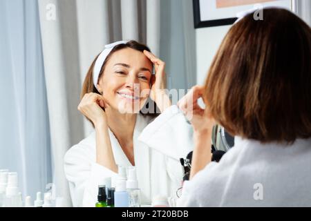 Femme souriante touchant le visage portant housecoat avant miroir Banque D'Images