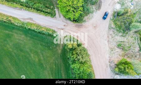 Une superbe perspective aérienne capturant une voiture noire conduisant sur un chemin de terre pittoresque entouré d'arbres verdoyants, de prairies et de champs, un voyage tranquille à travers la nature. Vue aérienne de Black car sur la route de terre. Photo de haute qualité Banque D'Images