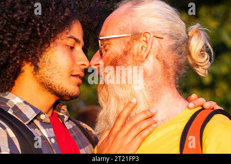 Un moment tendre et passionné capturé comme un homme arabe gay millénaire attrayant avec de longs cheveux bouclés et son amant masculin caucasien plus âgé avec de longs cheveux gris et un regard de barbe l'un sur l'autre, sur le point de partager un baiser passionné dans l'étreinte chaude du soleil couchant. Moment intime de coucher du soleil, baiser de couple gay. Photo de haute qualité Banque D'Images