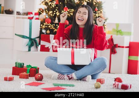 Photo de la mignonne dame chanceuse habillée tricotée pull de noël levant les poings devenant présent à l'intérieur de la chambre de la maison Banque D'Images