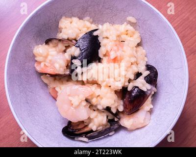Risotto italien aux fruits de mer, riche et crémeux, orné de moules, crevettes et riz infusé au safran, présenté dans un bol blanc reposant sur une planche de bois. Banque D'Images