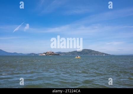 Vue panoramique de l'île d'Alcatraz avec un bateau flottant sur l'eau de l'océan ondulante contre le ciel bleu avec des nuages et des montagnes à San Francisco Banque D'Images