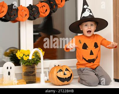 Concept Halloween. Un petit garçon joyeux et beau dans un chapeau de sorcier et un chandail orange est assis sur une table dans la cuisine sur le fond d'un Banque D'Images