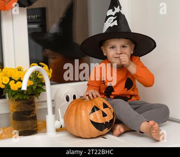 Concept Halloween. Un petit garçon joyeux et beau dans un chapeau de sorcier et un chandail orange est assis sur une table dans la cuisine sur le fond d'un Banque D'Images