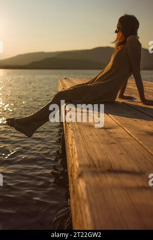 Une jeune femme caucasienne contemplative est assise seule sur une jetée au bord du lac, contemplant l'eau au coucher du soleil ou à l'aube, dans une atmosphère orange et vintage. Banque D'Images