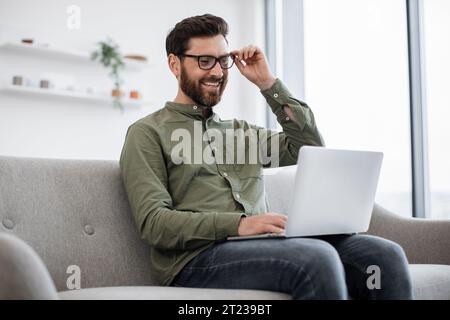 Pensive homme barbu calme portant des vêtements décontractés ajustant des lunettes tout en étant assis sur un canapé confortable et en utilisant un ordinateur portable moderne. Concept de gadgets, freelance Banque D'Images