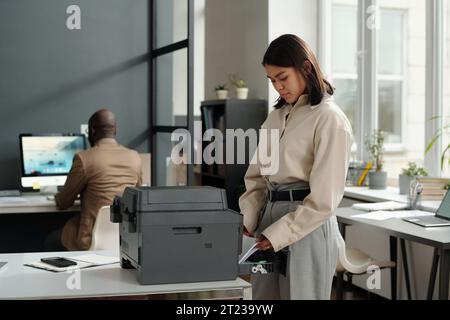 Jeune femme d'affaires brune en vêtements formalwear mettant des feuilles de papier dans le bac xerox tout en se tenant devant le copieur par son lieu de travail dans le bureau Banque D'Images