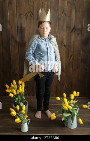 Portrait d'un garçon portant une couronne en carton et tenant une épée, avec des bouquets de tulipes jaunes debout à proximité. Cadre vertical. Regarde la caméra. Banque D'Images