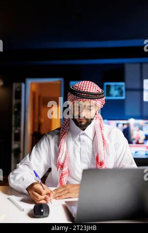 Un homme musulman professionnel utilise la technologie pour travailler efficacement au bureau, naviguer en ligne et écrire des notes à partir d'un ordinateur portable. Jeune étudiant arabe utilisant son cahier et son miniordinateur pour la recherche. Banque D'Images