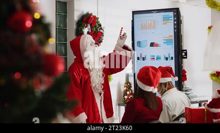 Manager habillé en Père Noël tenant une réunion d'équipe, montrant la stratégie de l'entreprise de l'année prochaine sur l'écran numérique. Chef d'équipe dans un bureau décoré festif parlant avec les employés Banque D'Images