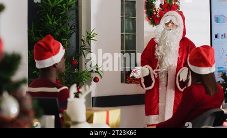 Manager habillé en Père Noël tenant une réunion d'équipe, présentant les prévisions de revenus financiers sur écran numérique. Chef d'équipe dans un bureau décoré festif motivant le personnel de l'entreprise Banque D'Images