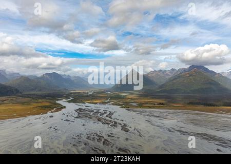 Une vue aérienne montre une large rivière tressée qui coule dans une vallée entourée de montagnes sous un ciel partiellement nuageux. Les multiples canaux de la rivière créent des motifs complexes à travers le paysage, avec l'eau apparaissant limoneux ou glaciaire en couleur. Le fond de la vallée est un mélange de zones herbeuses et de parcelles d'arbres, menant aux pentes boisées des montagnes. Le ciel est un mélange de nuages bleus et blancs, ajoutant de la profondeur et de la texture à la scène. Banque D'Images