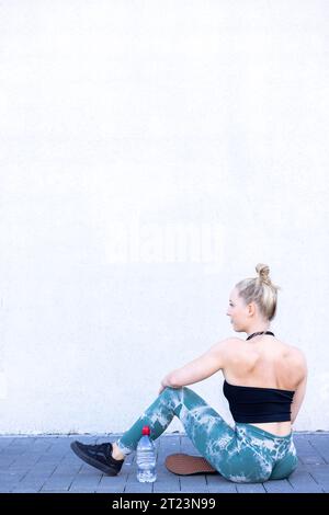 Une femme blonde sportive, son dos musclé à la caméra, est assise avec bonheur et joie après un entraînement en plein air, regardant par-dessus ses épaules. Elle tient une bouteille d'eau, incarnant un mode de vie sain et actif. Femme blonde sportive se reposant après un entraînement en plein air. Photo de haute qualité Banque D'Images