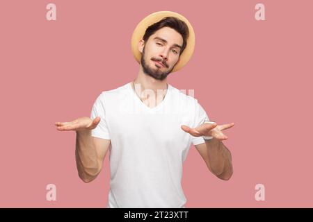 Portrait de bel homme barbu attrayant confiant en T-shirt blanc et chapeau debout regardant la caméra, montrant le geste cool rock, semble ludique. Studio intérieur tourné isolé sur fond rose. Banque D'Images