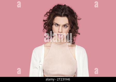 Portrait d'une folle femme folle avec les cheveux bouclés portant une tenue de style décontracté ayant une expression faciale stupide, grimacant, montrant de mauvaises manières. Studio intérieur tourné isolé sur fond rose. Banque D'Images