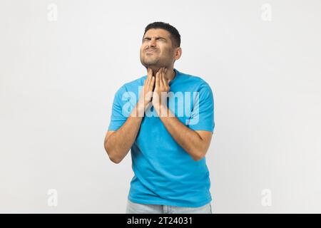 Portrait d'un homme malade triste et non rasé portant un T-shirt bleu se tenant debout touchant son cou douloureux, ayant mal à la gorge, fronçant le visage, étant très malade. Studio intérieur tourné isolé sur fond gris. Banque D'Images