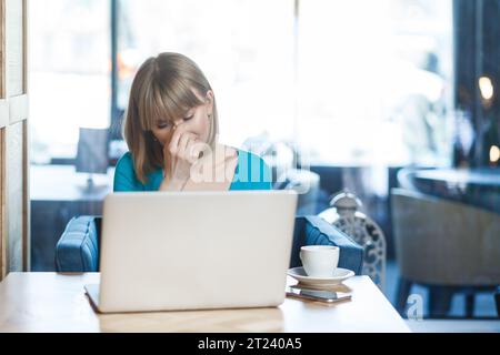 Portrait de triste désespoir bouleversé jeune femme aux cheveux blonds en chemise bleue travaillant sur ordinateur portable, pleurant, cachant son visage, ayant des problèmes avec son travail. Tourné à l'intérieur dans un café avec une grande fenêtre sur fond. Banque D'Images