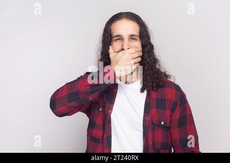 Portrait d'homme barbu avec de longs cheveux bouclés en chemise rouge à carreaux essaie de ne pas dire secret à ses amis, couvre la bouche tout en commérant. Studio intérieur tourné isolé sur fond gris. Banque D'Images