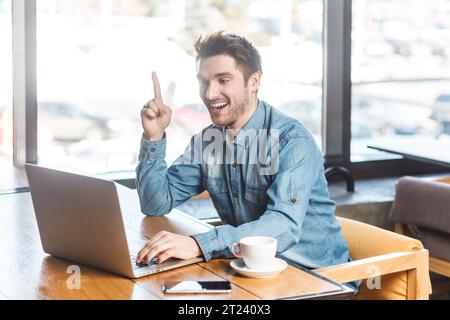 Portrait de jeune homme excité overjoyé travaillant sur ordinateur portable et levé le doigt vers le haut, ayant une bonne idée, a une réunion en ligne, discuter du projet. Prise de vue intérieure près de la grande fenêtre, fond de café. Banque D'Images