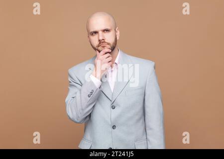 Laisse-moi y réfléchir. Portrait de pensif bel homme barbu chauve tient le menton et regarde pensivement loin essaie de résoudre le problème, portant une veste grise. Studio intérieur tourné isolé sur fond brun Banque D'Images