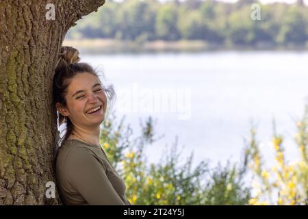 Découvrez la sérénité pure comme une charmante jeune brune millénaire embrasse la tranquillité d'une forêt verdoyante. Assise contre un arbre robuste, elle sourit chaleureusement, se prélassant dans la beauté de la nature. Son regard rencontre la caméra, transmettant un sentiment de paix au bord du lac de la forêt bleue sereine en cette délicieuse journée d'été. Sérénité au milieu de la nature : Smiling Millennial in Forest Escape. Photo de haute qualité Banque D'Images