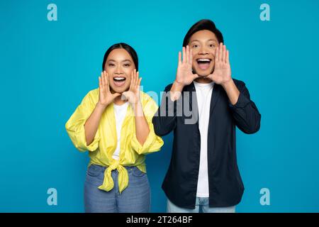 Heureux couple asiatique dans décontracté mettant les mains à la bouche, hurlant, isolé sur fond de studio bleu Banque D'Images
