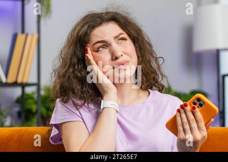 Portrait de femme fatiguée freelance assis sur le canapé utiliser smartphone, souffrant de maux de tête problème tension et migraine, stress. Fille adulte envoie des messages, fait des achats en ligne à l'intérieur dans la chambre à la maison Banque D'Images