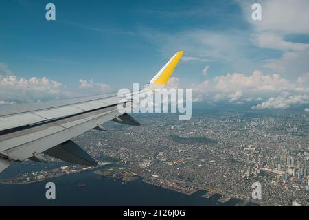 Wing from window of airplane flying over the city of Manila. Aerial view of Capital of Philippines. Manila bay and buildings below, clouds in blue sky. Banque D'Images