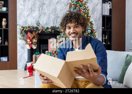 Portrait de l'homme heureux à la maison à Noël, homme hispanique tenant le cadeau de boîte aux lettres en carton, heureux reçu le produit de la boutique en ligne à l'heure, assis dans le salon près de l'arbre de Noël. Fête du nouvel an. Banque D'Images