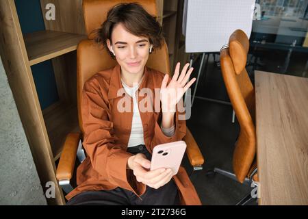 Portrait de femme disant bonjour, parlant à smartphone, chat vidéo, ayant une réunion en ligne dans son bureau, en utilisant le téléphone portable pour la conversation Banque D'Images