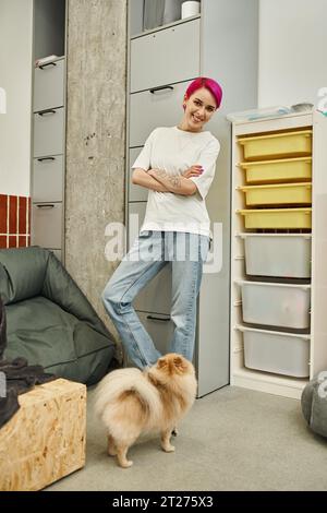 soignant tatoué et aux cheveux violets avec les bras pliés souriant à la caméra près de mignon doggy dans l'hôtel pour animaux de compagnie Banque D'Images