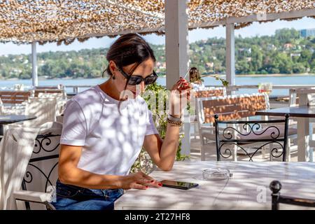 Portrait d'une jeune femme élégante fumant une cigarette assise à l'extérieur sur la terrasse du café au bord de la rivière. Habitude malsaine. Banque D'Images